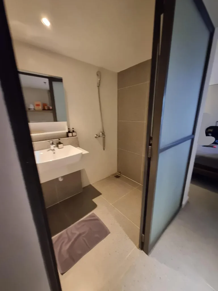 The bathroom of a fully furnished hotel room in Southeast Asia. The image shows a "wet bathroom", which is the style in Southeast Asia in which there is no divider between the shower and sink & toilet. The flooring is tile, and water from the shower can splash anywhere within the bathroom.