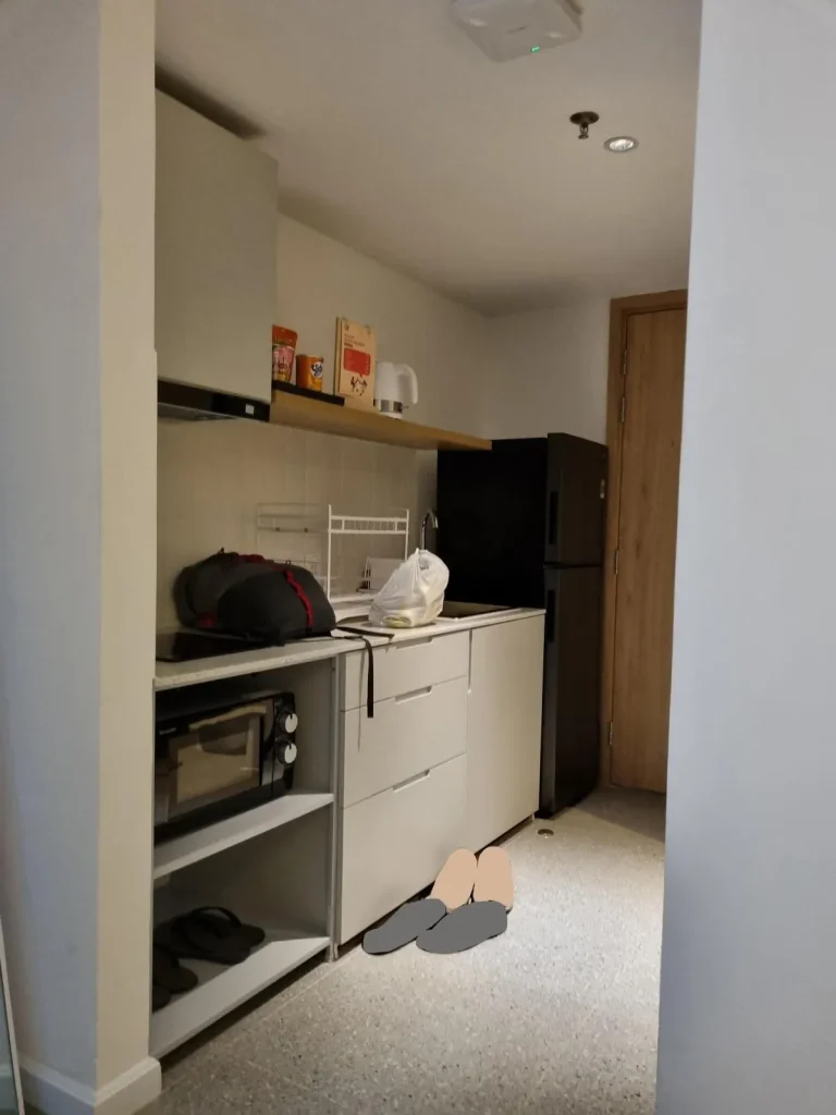 The kitchen area of a fully furnished hotel room in Southeast Asia. The image shows a toaster microwave, refrigerator, hot water kettle, stove and ventilation, and storage drawers with utensiles and pots & pans inside.