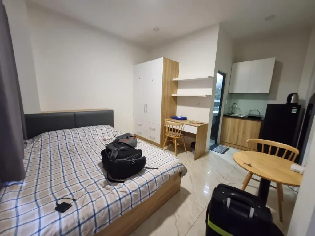 A fully furnished studio room rented monthly in Southeast Asia. The image shows a queen-sized bed with all sheets, a small round table, a wardrobe, shelf, table mounted to the wall, chairs, small kitchen & refigerator, and the bathroom door in the background. Not shown is the split-duct airconditioning unit. There is a window with curtains next to the bed.