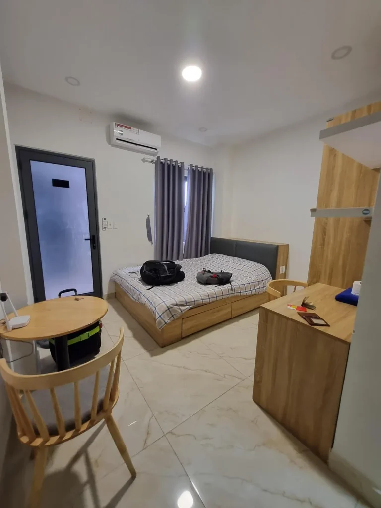 A fully furnished studio room rented monthly in Southeast Asia. The image shows the front door that is mostly a window with opaque film on it. Also shown a queen-sized bed with all sheets, a small round table with chair, table mounted to the wall with a chair, ceiling lighting, and the split-duct airconditioning unit. There is a window with curtains next to the bed.