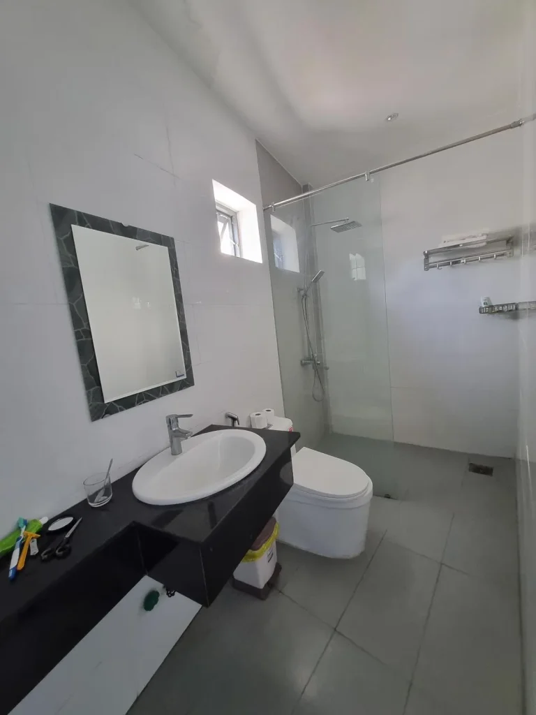 The bathroom of a furnished hotel room in Southeast Asia without a kitchen. It has a partial divider between the shower and toilet & sink area. A "wet bathroom" is the most common style in Southeast Asia in which there is no divider between the shower and sink & toilet. The flooring is tile, and water from the shower can splash anywhere within the bathroom.