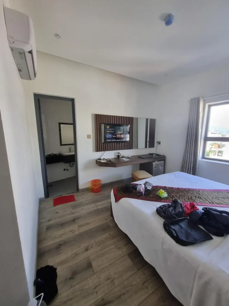 A furnished hotel room in Southeast Asia without a kitchen. The image shows a split-duct airconditioning unit, bathroom entrance, a queen-sized bed with all sheets, a window with draperies, a table, refrigerator, television, nightstand, and trashcan. The tenant has belongins laying on the bed, nightstand, and floor.