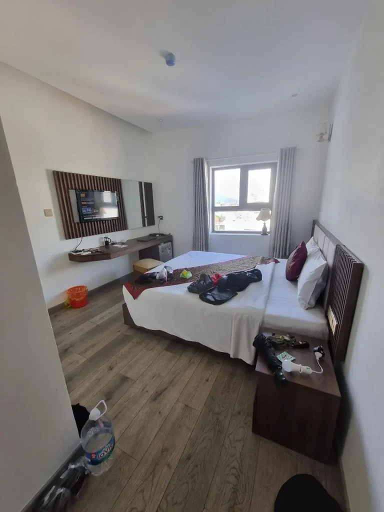 A furnished hotel room in Southeast Asia without a kitchen. The image shows a queen-sized bed with all sheets, a window with draperies, a table, refrigerator, television, nightstand, and trashcan. The tenant has belongins laying on the bed, nightstand, and floor.