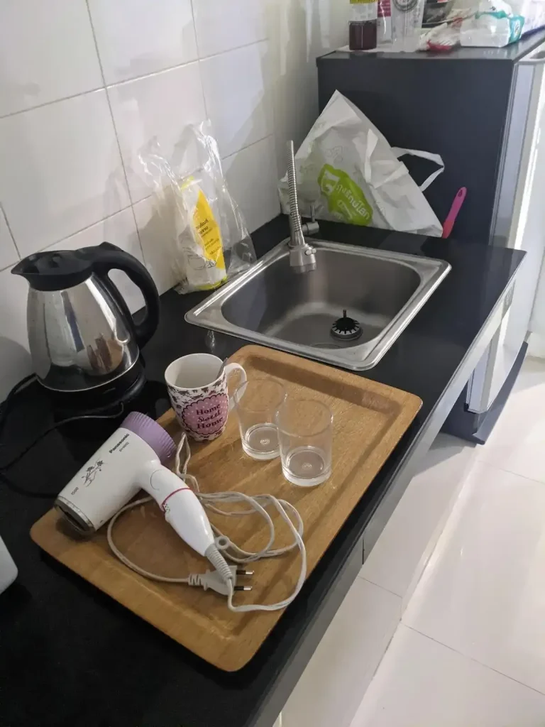 An image showing part of the kitchen in a furnished one bedroom apartment that is rented monthly in Southeast Asia. The kitchen is it's own, separate, room. In this kitchen area few things furnished by the apartment complex including a water boiling kettle, cups, a tray, and a hair dryer.