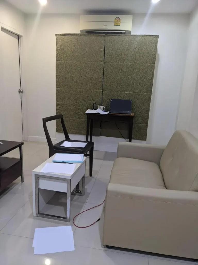 A furnished one bedroom apartment that is rented monthly in Southeast Asia. This image is of the family room eqiupped with a small leather couch, small coffee table, and a window along the wall to the side. In front of the window is a desk with a chair with a laptop and coffee mug on it. A split-duct airconditioning unit above the window. The floor is tile, which is very common in Southeast Asia.