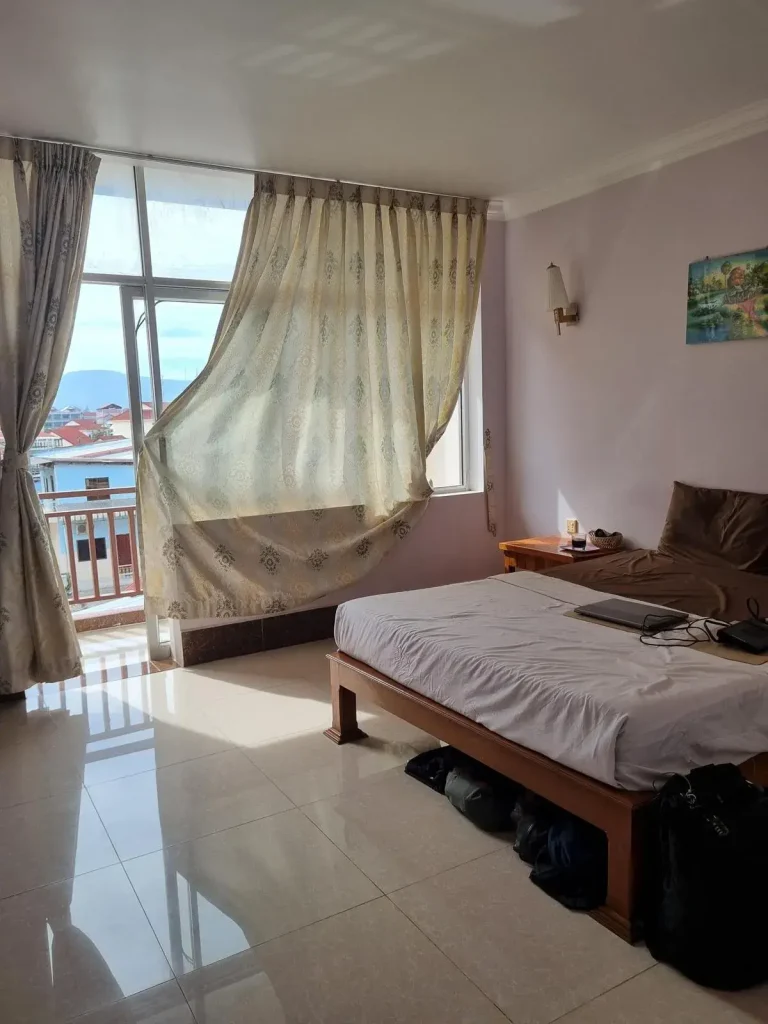 A furnished studio room rented monthly in Southeast Asia. The image shows a queen-sized bed with all sheets, and the large windows with curtains and a glass door opening to a balcony.