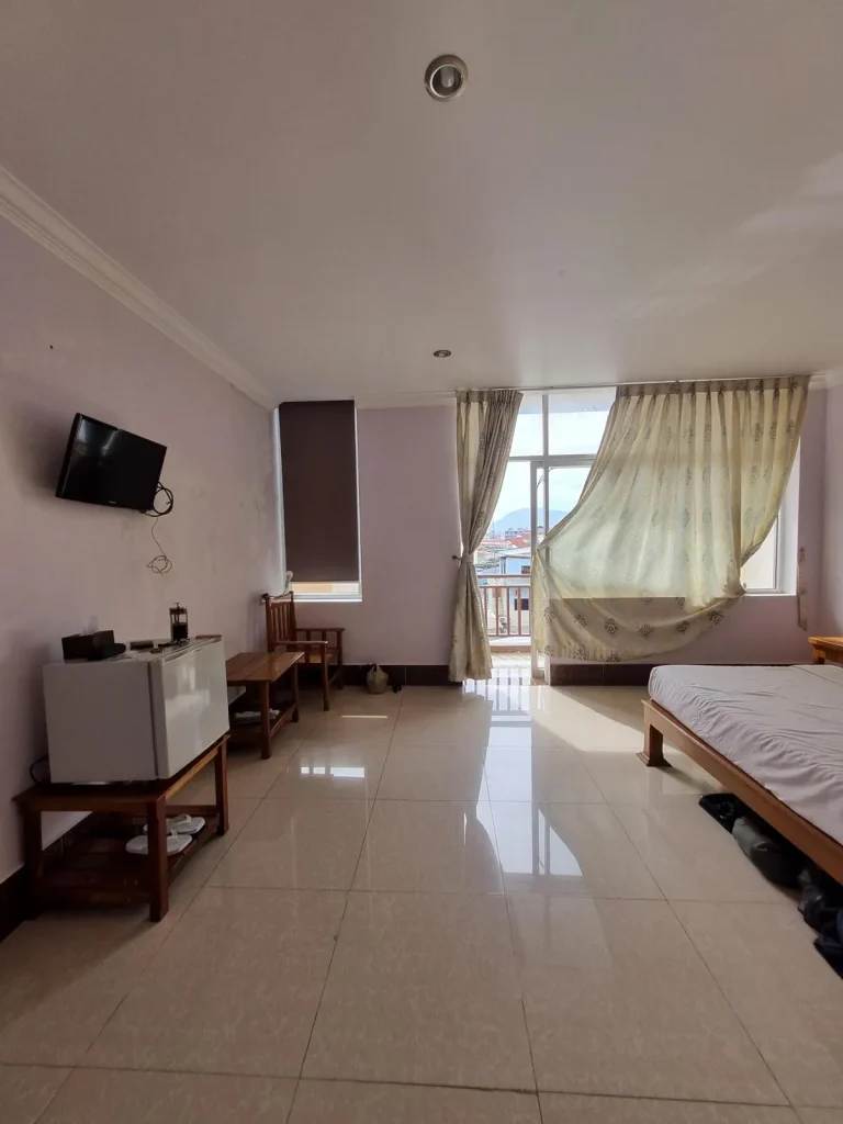A furnished studio room rented monthly in Southeast Asia. The image is at the entrance of the room getting a view of the left-hand side of the room that has a small refrigerator, a small television, a small table and a chair. On the back-side wall, there is a window near the chair, and large windows with curtains and a glass door opening to a balcony. Not shown is the split-duct airconditioning unit.