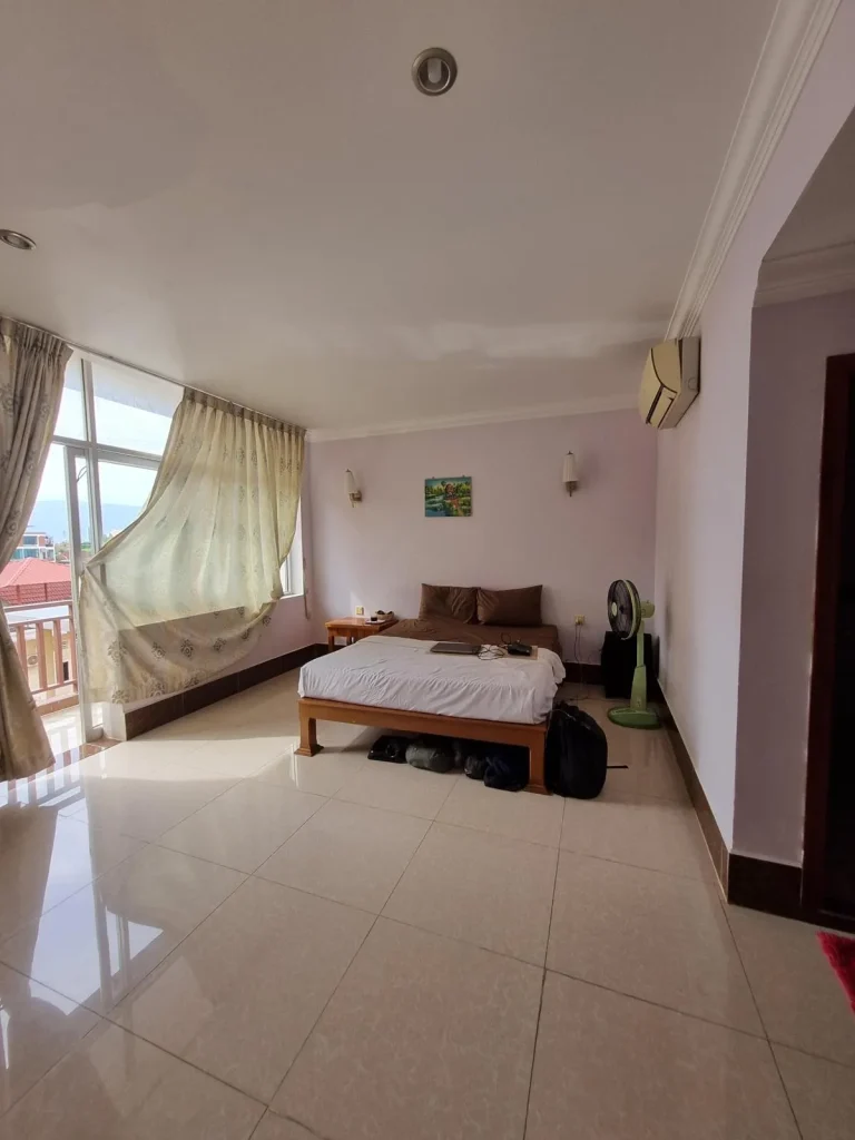 A furnished studio room rented monthly in Southeast Asia. The image shows the right-hand side wall of the room. This includes a queen-sized bed with all sheets, a floor fan to cool down, a split duct airconditioning unit, and nightstand. To the left - the back-side of the room- are large windows with curtains and a glass door opening to a balcony.