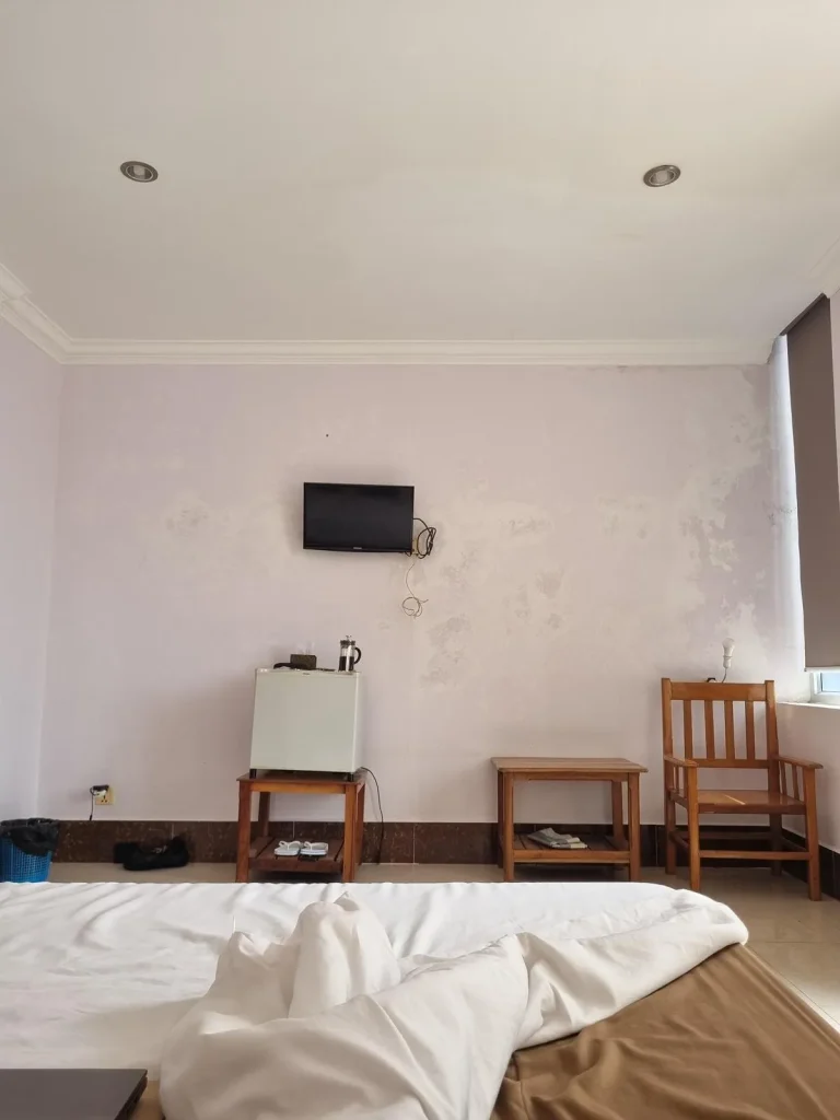 A furnished studio room rented monthly in Southeast Asia. The image shows the left-hand side wall of the room. This includes the edge of the bed. Along the wall is a small refrigerator, two small tables, a chair, a small television, and a window. The wall paint is peeling off a bit.