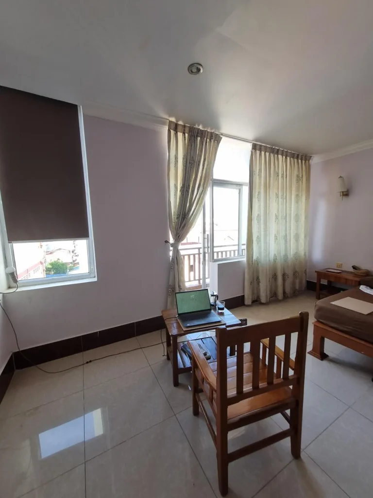 A furnished studio room rented monthly in Southeast Asia. The image shows the back side wall of the room. A window to the left, and to the right are large windows with curtains and a glass door opening to a balcony. The tenant has a setup in the middle of the room with a laptop on the small table, and a chair pulled up next to it facing outside, looking out the large windows.