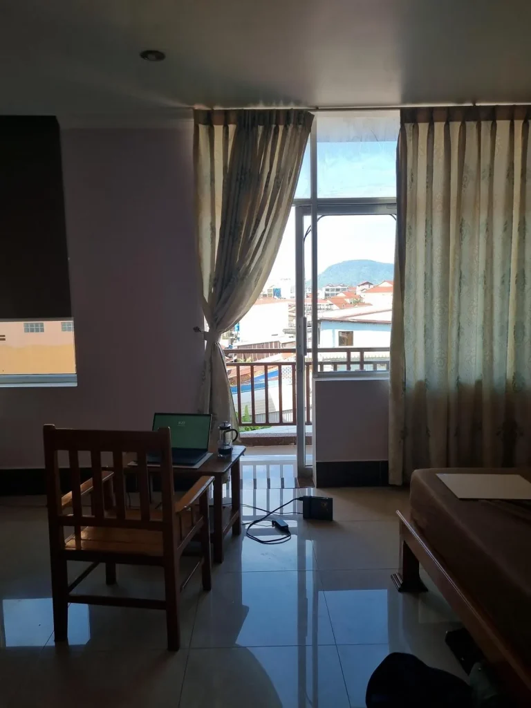 A furnished studio room rented monthly in Southeast Asia. The image shows the back side wall of the room. A window to the left, and to the right are large windows with curtains and a glass door opening to a balcony. The tenant has a setup in the middle of the room with a laptop on the small table, and a chair pulled up next to it facing outside, looking out the large windows.