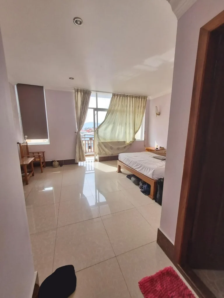 A furnished studio room rented monthly in Southeast Asia. The image is at the entrance of the room with the bathroom the the right, a queen-sized bed with all sheets, windows with curtains and a glass door opening to a balcony, another window to the left, and a small table with a chair. Not shown is the split-duct airconditioning unit.