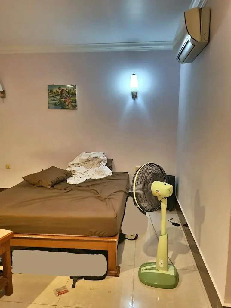 A furnished studio room rented monthly in Southeast Asia. The image shows a close-up of the bed, portable floor air fan, and a split-duct airconditioning unit.