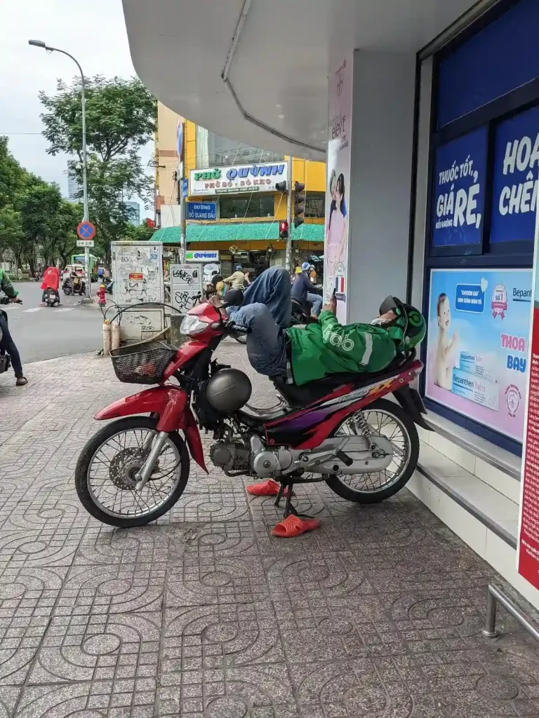 On a sidewalk, a driver of a motorbike taxi is laying on his parked motorbike, relaxing. His back is on the seat, and his feet are on the front handle bars in a fetal position. He is employed through the Grab ride hailing company, and is wearing Grab-branded jacket. His helmet is Gojek branded, a ride hailing company similar to Grab.