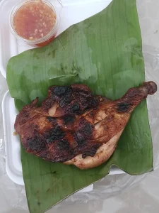Grilled chicken chop with a side of garlic sauce in Cambodia. The chicken is laid on a banana leaf within a styrofoam take-out container.