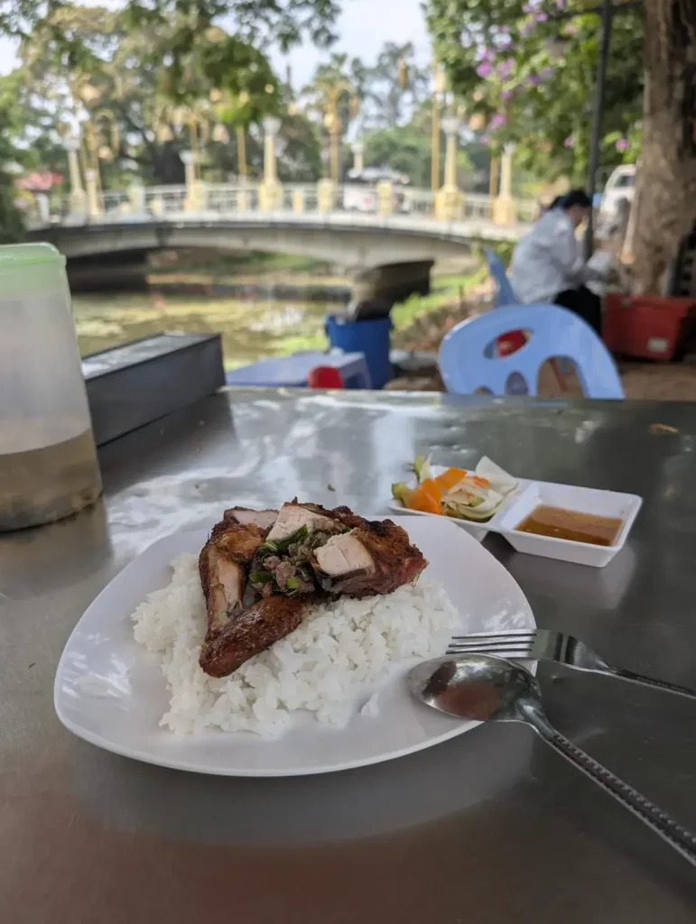 Outdoor table and seating along the sidewalk, at an outdoor food stall, with a plate of chopped grilled chicken on rice in Southeast Asia. The dish has pickled vegetables as a condiment, sauce, and a jug of tea on the table for customers. The background shows that the table is also along a small river with a bridge crossing from one side to the other. Trees abound.