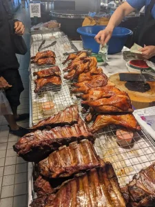 A counter of grilled meat at a food market in Thailand. Grilled pork ribs, chicken chops, and whole chickens are for same. The seller has a meat clever and chopping board to chop up the meat as you desire. A customer is purchasing something in this image, exchanging cash with the seller.