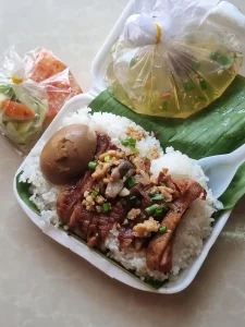 A portion of sliced grilled pork on rice with a boiled egg, pickled vegetables, and a packet of sauce with red chillies inside. The dish comes with a soup. This dish is from Cambodia, but closely resembles a Vietnamese version.