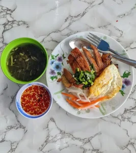 On a granite table is a plate of chopped grilled pork on rice with a fried egg, pickled carrot, and a small bowl of sauce with red chillies inside. The dish comes with a soup. This dish is in Vietnam.