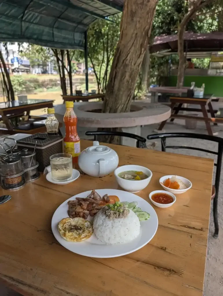 Outdoor table and seating with overhead covering along the road in Southeast Asia. On the customer's table is a plate of chopped grilled pork on rice with a fried egg, sliced cucumber and tomato. The dish comes with a soup, and pickled vegetables as a condiment, sauce, and a pot of tea on the table for customers. The background shows that the table is also along a small river. There is a tree within the seating area that pokes through the overhead cover, and several small trees beyond the covering's structure.