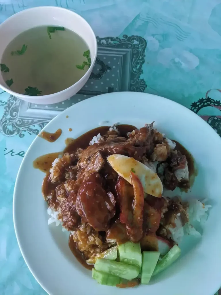 A dish in Laos called grilled red pork rice, and it is just exactly that. It comes with sliced cucumber on the side, sliced boiled egg, a few slices of Chinese sweet sausage, and a brown sauce that covers the entire dish. A simple brothy soup is included.