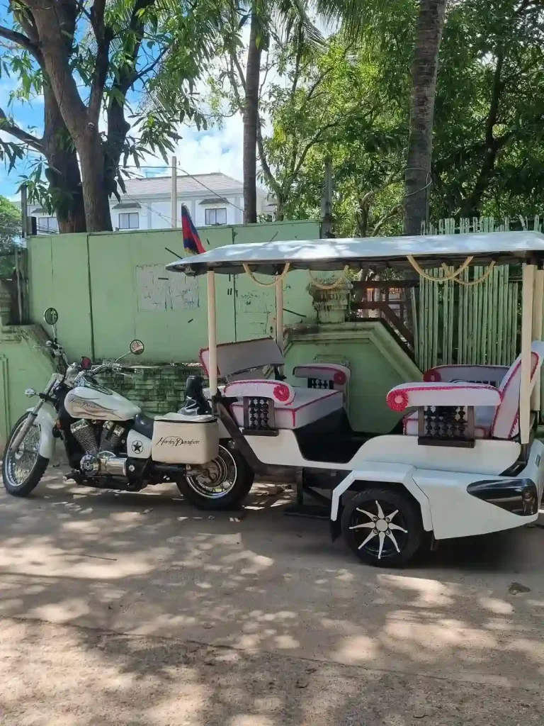 A fancy remorque (French word for wagon) taxi that is towed by a Harely Davidson in Cambodia. It is parked. A remorque "wagon" consists of a motorbike that is towing a two-wheeled wagon with seating and a canopy. It has two bench seats - one looking forward, and another one looking backward. Each bench seat fits two people, so four people total can ride in this taxi. This attached wagon-style transportation is unique to Cambodia - it can be used as a family vehicle, a taxi, and sometimes even for moving goods. The motorbike is a Harely Davidson motorcycle in this image.