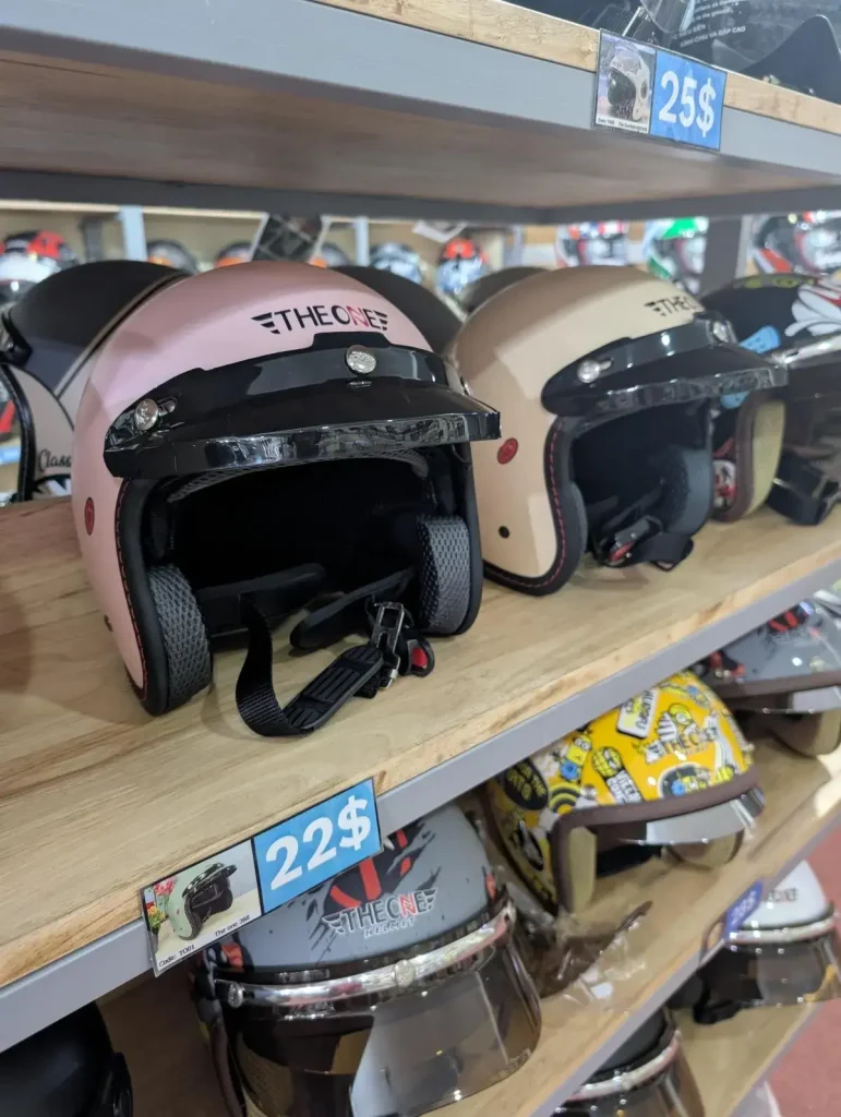 Helmets of various styles at a helmet shop in Southeast Asia. The helmet in the image have a face shield feature that can be pulled downward (to use the face shield) or upward (to get rid of the shield). There is no chin coverage (i.e. it is not a full-face-covered helmet). Price is twenty-two US dollars.