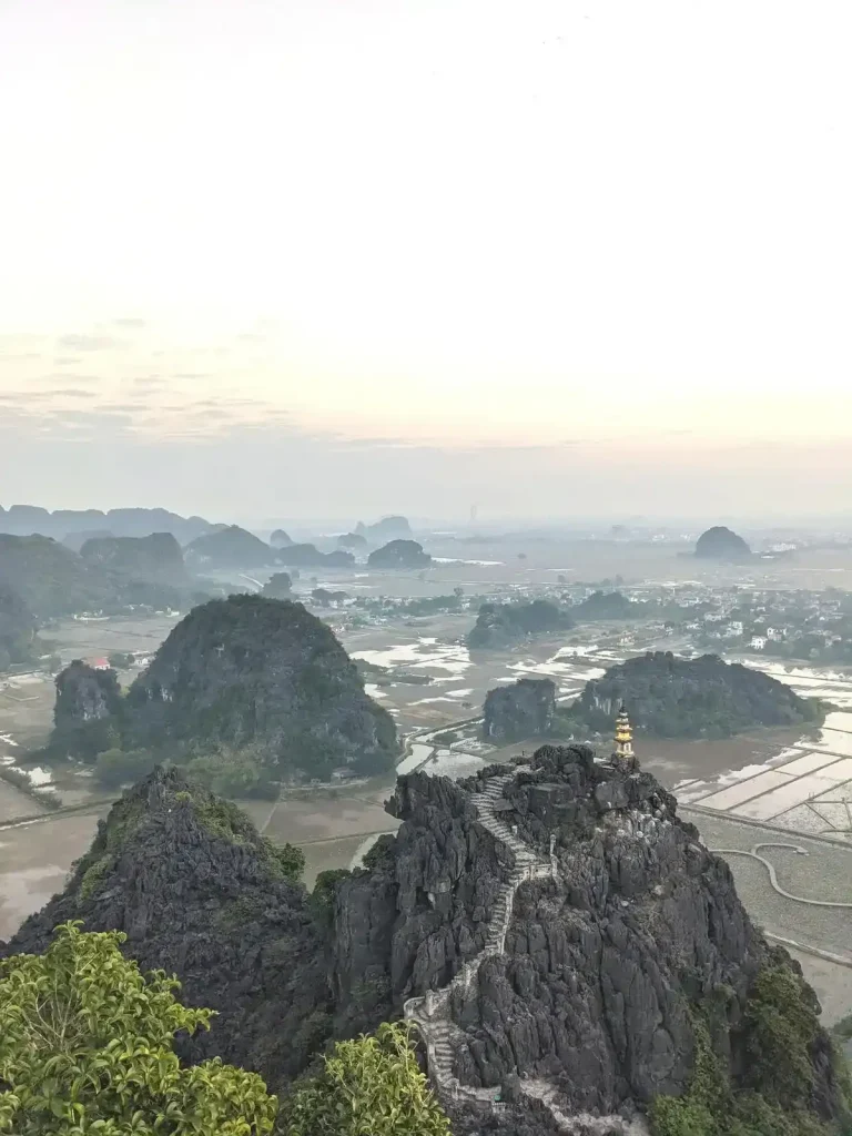 An overlook in Ninh Binh, Vietnam of several steep limestone mountains that pop-up throughout the distant land like moles (an animal) popping their heads out of the ground. The closest mountain has a hiking trail carved into the stone, leading to a small golden-painted temple at it's peak.