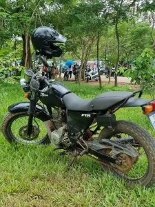 A Honda FTR motorcycle that is fully manual (uses a hand clutch lever, in part, to change gears). It is parked in a grassy area with trees. The engine size is larger (223 cc) than a typical SE Asian motorbike (125 cc). Also, it has bulkier tires, which is good for rough terrain such as bumpy dirt roads.