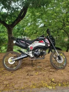 A Honda Grom manual motorbike in Cambodia. It is retro-fitted with dirt bike wheels (rims and tires). It is parked in a small patch of ground - no grass just dirt - with trees in the background not far away. The trees make a nice backdrop for the image.