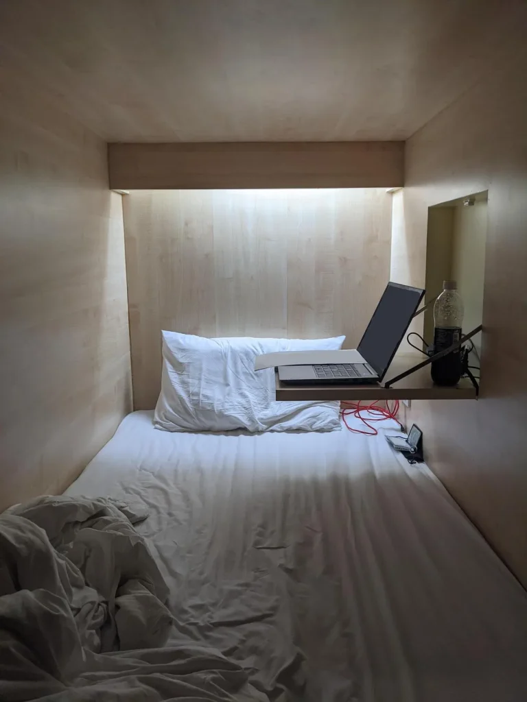 A private bed contain within it's own walled pod in a dorm room at a hostel in Southeast Asia. The pod is the exact width of a twin mattress. It's entrance has a curtain. Inside is a mattress, pillow, sheets, and a fold-out table that is built into the wall.