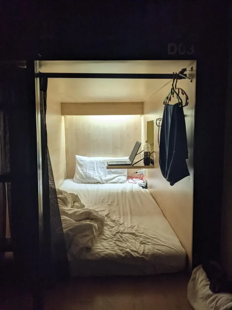 A private bed contain within it's own walled pod in a dorm room at a hostel in Southeast Asia. The pod is the exact width of a twin mattress. It's entrance has a curtain. Inside is a mattress, pillow, sheets, and a fold-out table that is built into the wall.
