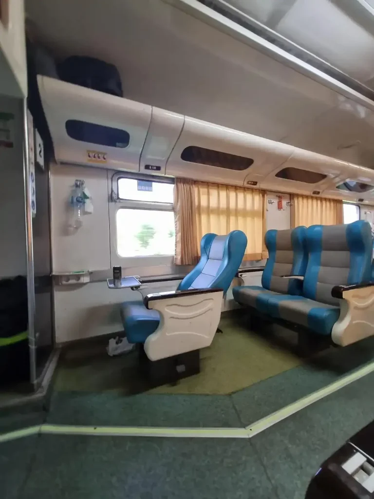 Indonesia train first class seating. There are 2 seats side-by-side with lots of leg room and lots of padding. The seats include a fold-out table for food on board, arm rests, and ability to recline backwards. Baggage storage is above the seating. Windows have curtains. The layout is 2 seats side-by-side, a middle aisle walk way, and another 2 seats. All seats are facing forward.