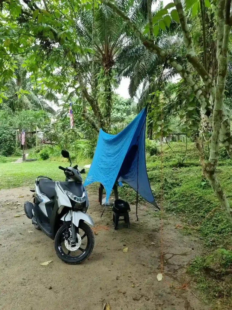 Hammock camping with a tarp covering the hammock at a camping park in Malaysia with jungle surroundings. The camping spot is cleared-out with a dirt ground and no plants invading the space. A motorbike is parked next to the hammock, presumably because the camper is on an adventure.