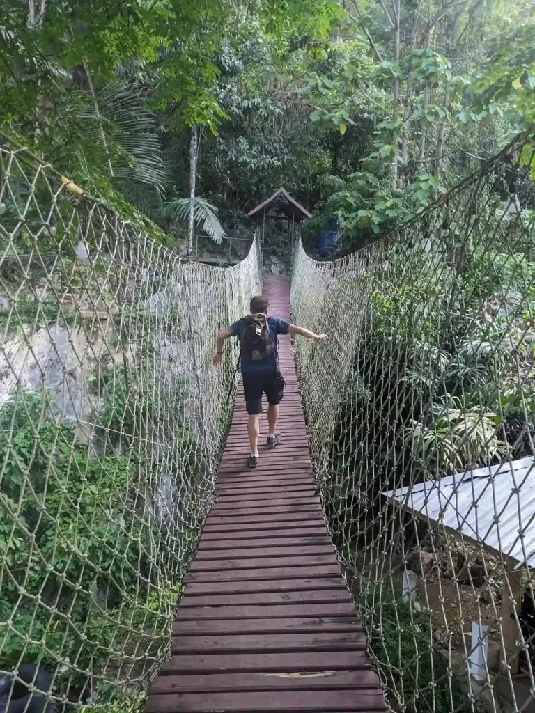 A hiker crossing a suspension bridge while hiking in a mountainous jungle region of Laos.