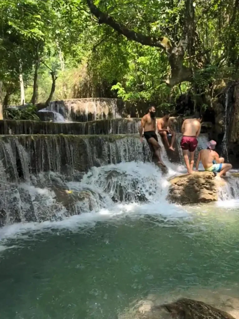 Four foreign men in swim trunks hanging out at a section within Kuangsi waterfall in Luang Pragang, Laos. This waterfall section has three tiers, each tier with a pool and then a drop-off that the water flows over to the next tier. The men are standing on a rock at the lower tier. At this lower tier is a pool of water good for simming in. It is a hot and bright day, but many tree branches cover this waterfall area, providing shade and a beautiful view.