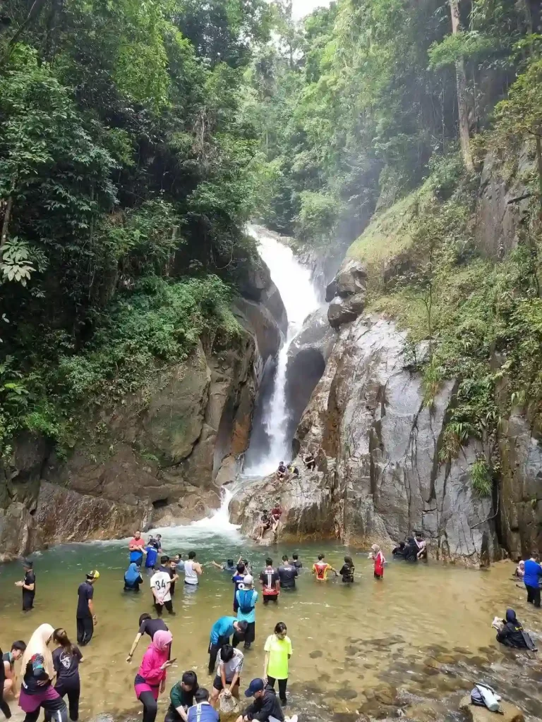 A large waterfall that rushes down a rocky crevice within the jungle. The water splashes onto a rock, then flows into a water pit area, where many (about thirty) people are playing in the water. This is located in Malaysia, a muslim country, and therefor people dress modestly while simming. Men and women doe not show skin, and women wear hijabs.