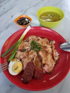A Laotian pork dish. Aside from the cut of pork mean, it includes two slices of Chinese sausage, half of a boiled egg, a stick of green onions (i.e. scallions), a soy-based sauce on the side, and a simple soup. Utensils used are spoon and a fork to push the food onto the spoon.