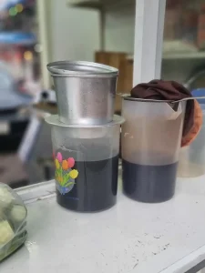 A large Phin coffee brewing filter (pronounced "feen" as in the ending of the wording "caffeine") resting on top of a plastic jug, brewing coffee. This large Phin brews an entire pot of coffee, instead of individual cup-sizes. This coffee filter is filled with ground coffee beans, and hot water is added inside. It uses gravity to slowly percolate (drip) hot water through the coffee grounds. At the bottom of this filter, the hot coffee drips out into a cup. This brewing method was introduced to Vietnam and Cambodia by the French during the colonial occupation time period.
