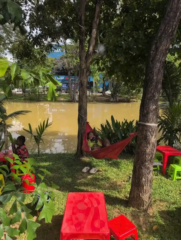 A woman is laying in a hammock that is suspended between two trees in the grass along the river in Siem Reap, Cambodia. She is watching videos on her cell phone, passing time until a customer stops at her food stall to eat.