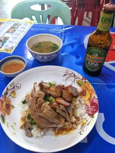 A pork dish in Laos consisting of pork leg, grilled red pork, and crispy pork on rice with a little bit of brown sauce added ontop. It comes with sliced cucumber and a garlic sauce to be added. A simple brothy soup is included. A Beer Lao IPA beer is alongside the dish.