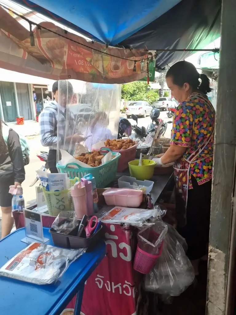 A food stall along a small road in Thailand serving food for people on foot and/or parked motorbike (scooter). The female seller is preparing a dish made with fried chicken tenders on rice. The chicken tenders are mixed with various sauces or spicies per the customers' choosing.