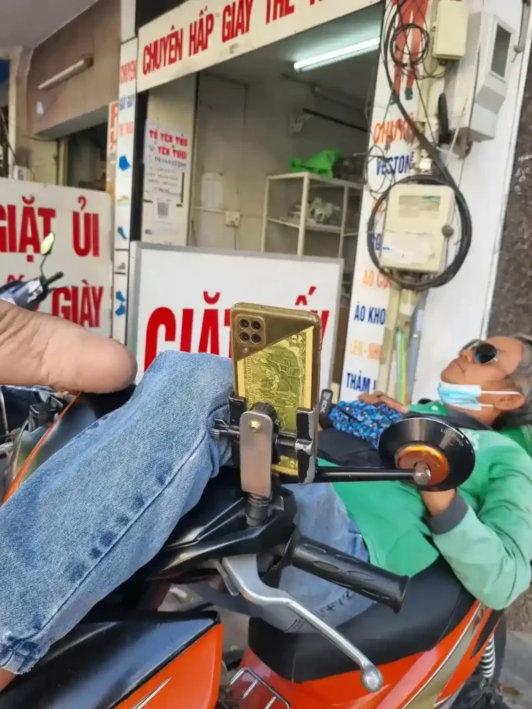 A local man relaxing on his parked motorbike in Vietnam. He is laying down. His back is on the seat, and his legs are wrapped over the handlebars. He has sunglasses on, and appears to be taking a nap. His phone is mounted on the handlebar, and the phone case has a two US dollar bill sandwhiched between his clear phone case and phone. This two dollar bill is considered for good luck in Vietnam.