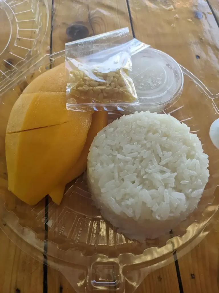 A serving of mango sticky rice, a popular desert in Thailand. It consists of a ripe and juicy mango that is fully pealed and ready to eat. There is a portion of sticky rice, a strain of rice that is litteraly sticky. Finally, there is some sweet coconut cream and a nut-like topping (it does not taste like nuts, though - not sure what to call it). Each ingredient is standalone and packaged for take-out (aka "to-go" or "take-away"). The person eating this will pour the coconut cream onto the mango and/or sticky rice, and the toping, and eat everything mixed together.
