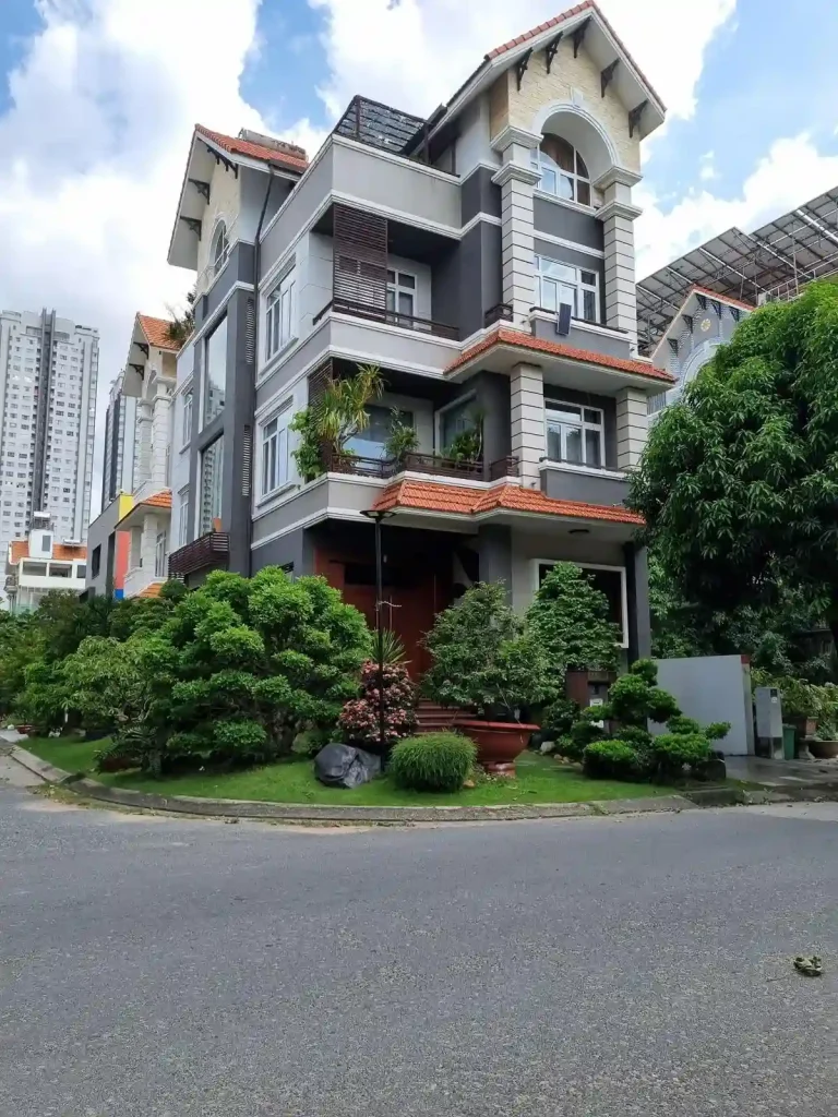A modern concrete four story home in Southeast Asia with a small grass lawn and various plants. This home is located on a small corner lot. The second and third floors each have a balcony with doors and windows. The fourth floor is half inside, and half a rooftop without covering for rain and sunlight.