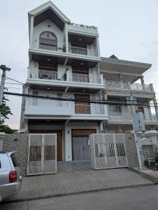 A modern concrete four story home in Southeast Asia. The ground floor has a gate and a small area for parking. The front door is not far from the street. The remaining floors each have a full-sized balcony with doors and windows. There is also a rooftop with some plants, but not covering for rain and sunlight.