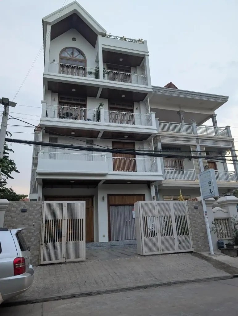 A modern concrete four story home in Southeast Asia. The ground floor has a gate and a small area for parking. The front door is not far from the street. The remaining floors each have a full-sized balcony with doors and windows. There is also a rooftop with some plants, but not covering for rain and sunlight.