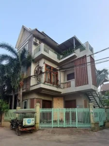 A modern concrete three story home in Southeast Asia. The ground floor has a gate and a small area for parking. The front door is not far from the street. There is also a rooftop with some plants.
