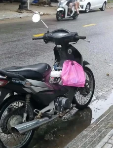 A plastic bag with it's handles hooked onto a hook accessory that is mounted on the dashboard area of a motorbike. The hook accessory gives riders a place to hold things that they purchas, as the lack much storage space on a motorbike.