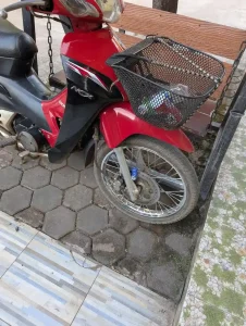 A basket with wire mesh mounted on the front of a motorbike in Southeast Asia. The basket is elevated above the front wheel, and has bungee chords on it to hold items in place. Inside is a water bottle and a can of Birdee-brand coffee. There is also a wheel lock on the disk brake of the front wheel to prevent theft while the motorbike is parked.